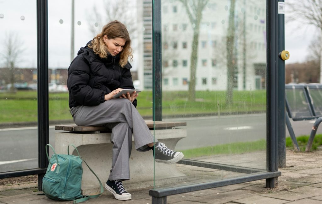 A young woman sitting at a glass bus stop looking at an electronic tablet