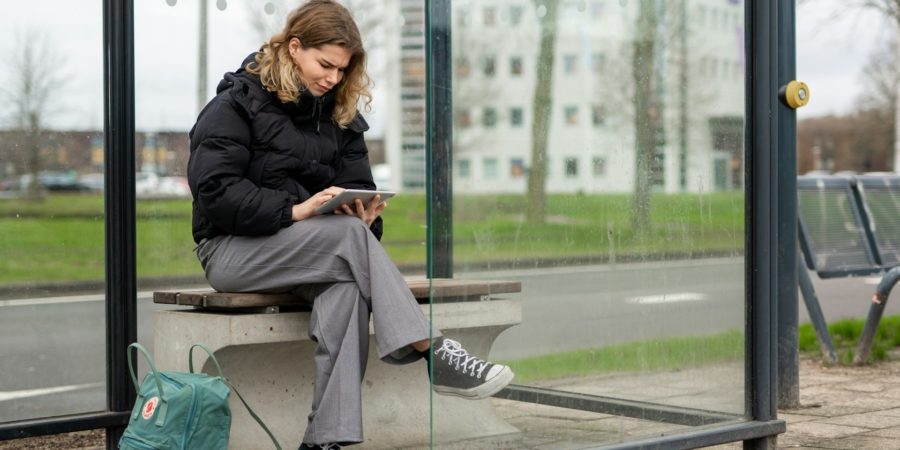 A young woman sitting at a glass bus stop looking at an electronic tablet